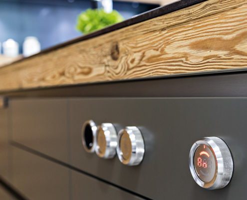 Baumann Küchen - Detailansicht von Drehreglern eines Herdes mit Edelstahl und Digitalanzeige auf dunkler Küchenfront und Echtholz Arbeitsplatte.