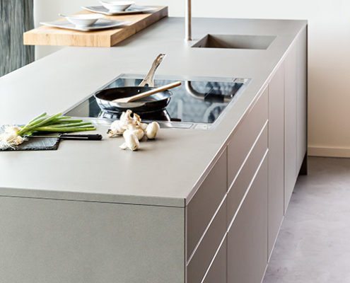 Baumann Küche - Seitenansicht einer hochwertigen Kochinsel in modernem grauen Design, mit Edelstahl Spülbecken, Kochfeld und Echtholz Servierfläche.