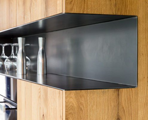 Küchen Baumann - Seitliche Detailaufnahme eines in die Küchenfront integrierten Regalfachs. Die Küchenfront besteht aus Echtholz und das Fach aus dunklem Metall.
