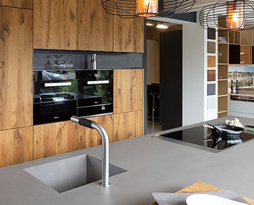 Baumann Küchen - Ansicht einer modernen Küche mit einer Kochinsel in grau mit Edelstahl Spülbecken und Kochfeld. Die Küchenfronten sind aus Echtholz und hochwertige schwarze Öfen sind integriert.