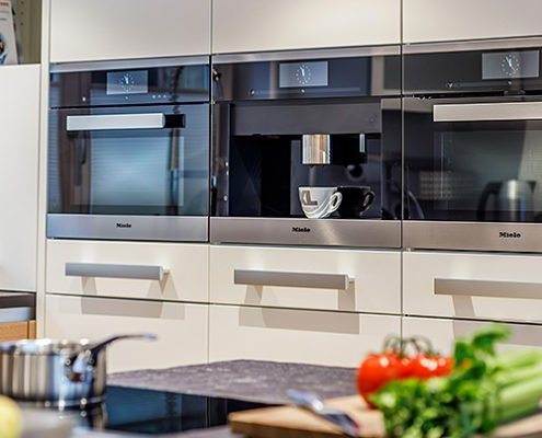 Küchenstudio Baumann - Blick in eine moderne Küche mit weißen Küchenfronten und breiten Griffen aus Edelstahl. Auf Augenhöhe drei Elektroeinbaugeräte von Miele, zwei schwarze Backöfen und ein Kaffeevollautomat.