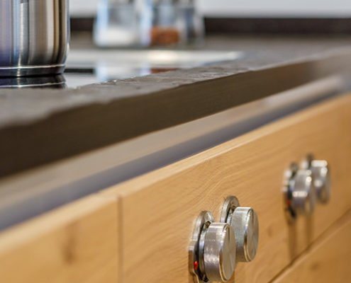 Küchen Baumann - Detailaufnahme von Einstellreglern eines Elektro-Herdes. Die Regler sind aus Edelstahl und auf einer Echtholz Küchenfront montiert. Die Arbeitsplatte besteht aus schwarzen, strukturierten Naturstein.