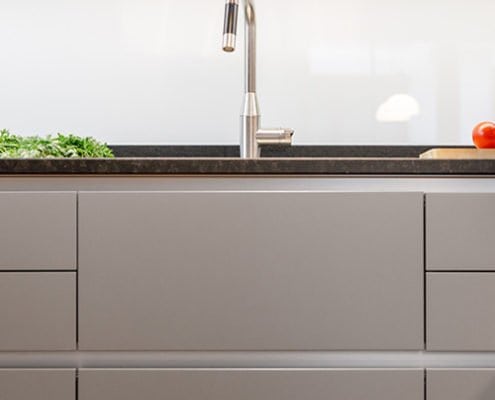 Baumann Küchen - Frontansicht von grauen Küchenfronten mit einer dunklen Arbeitsplatte aus Granit und einem Wasserhahn im Gastro Stil.