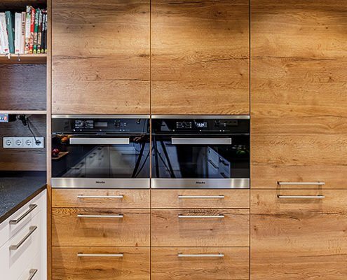 Küchenstudio Baumann - Frontansicht einer Küchenfront aus Echtholz, mit schmalen Edelstahl Griffen und zwei modernen, integrierten Elektrogeräten von Miele auf Brusthöhe.