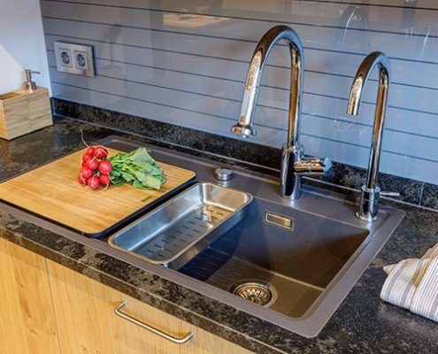 Baumann Küchenstudio - Detailansicht eines modernen Spülbeckens aus Edelstahl, mit zwei gebogenen Wasserhähnen, integriert in einer Arbeitsplatte aus Naturstein.